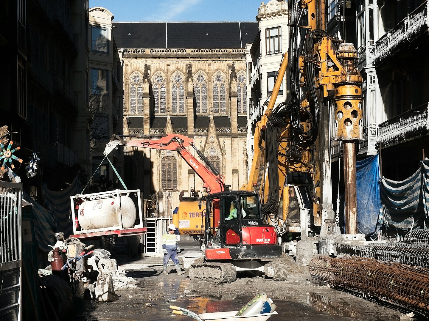 Operaio edile supervisiona macchinari pesanti in un cantiere urbano fangoso.